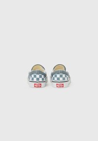 Zapatos slip-on a cuadros en lona gris y blanca, con suela de goma blanca y acento de logo rojo. Textura suave, forma redondeada.