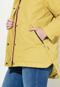 Gul vattentät jacka med dragkedja framtill, kontrasterande rosa dragkedja, sidofickor och en nedre fåll. Slät textur och avslappnad passform.