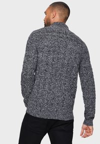 Strikket sweater i sort og hvid, med en tekstureret overflade, lange ærmer og høj krave. Ingen synlig branding. Bagfra vist.