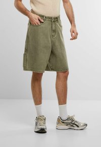 Shorts in denim verde oliva con una vestibilità ampia, dotati di tasche anteriori e tasca laterale cargo. Indossati con calze bianche fino alla caviglia e scarpe da ginnastica metallizzate.