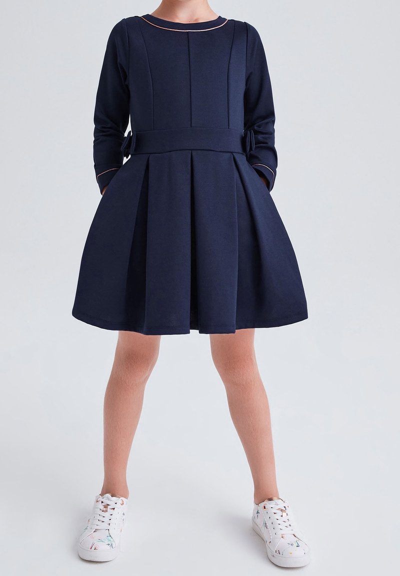 Enfant portant une robe plissée à manches longues bleu marine avec des poches et des baskets blanches à motifs floraux, sur un fond uni.