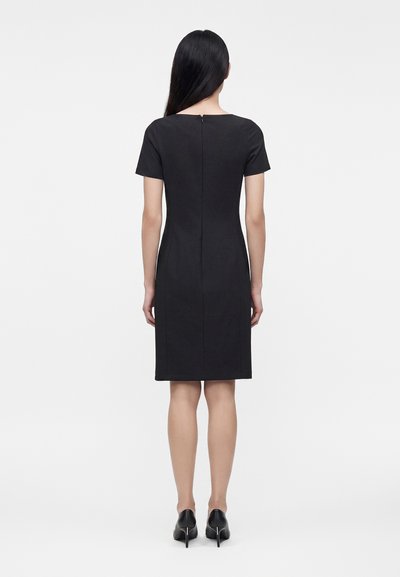 Femme portant une robe noire longueur genou avec manches courtes et des talons noirs, debout de dos devant un fond blanc uni.