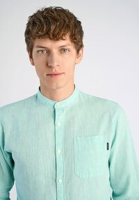 Vaalean mintunvihreä paita, jossa on kaulus, pitkät hihat ja rintatasku; siinä on hienovarainen rakenne ja nappikiinnitys.