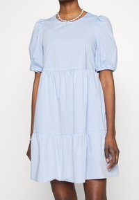 Světlé modré šaty s krátkým rukávem, nabíranými rukávy, nabíranými volány na lemu a kulatým výstřihem vyrobené z měkké, strukturované tkaniny.