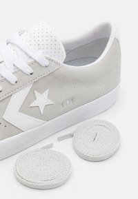 Ljusgrå mockasneakers med vit stjärnlogotyp, vita snören och två extra spiralformade vita skosnören på en vit bakgrund.