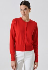 Frau in einer enganliegenden roten Knopfpulloverjacke und weißen Hosen, die vor einem schlichten hellen Hintergrund steht und leicht zur Seite blickt.