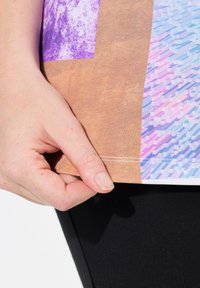 Camiseta multicolor con patrones en púrpura, marrón y azul. El primer plano muestra una mano sosteniendo el dobladillo, resaltando la tela y la costura.
