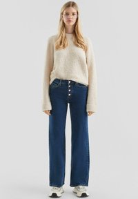 Licht beige, fluffy trui gecombineerd met high-waisted, wijde donkerblauwe jeans met vijfknopen sluiting, gedragen met witte sneakers.