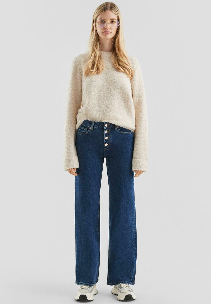 Licht beige, fluffy trui gecombineerd met high-waisted, wijde donkerblauwe jeans met vijfknopen sluiting, gedragen met witte sneakers.