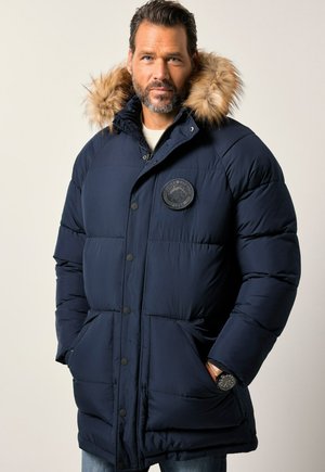 Hombre que lleva una chaqueta acolchada azul marino con capucha de piel sintética, con las manos en los bolsillos y un reloj de muñeca, de pie frente a un fondo claro y liso.