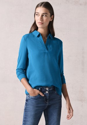 Blusa lunga turchese con colletto a V, realizzata in tessuto morbido, abbinata a jeans blu scuro con chiusura a doppio bottone.