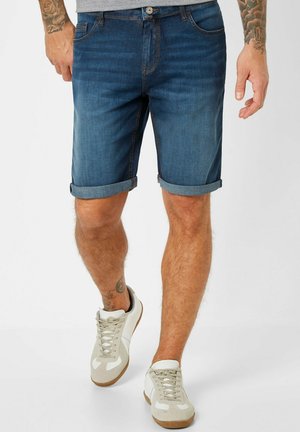 Mann trägt hochgekrempelte dunkelblaue Jeansshorts und weiße Sneakers, zeigt Tattoos an Armen und Knöcheln, läuft auf weißem Hintergrund.