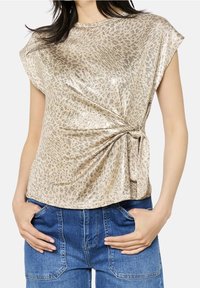 Metalický zlatý top s rukávky typu čepička, zdobený leopardím vzorem a uzlem vpředu, kombinovaný s modrými džínovými kalhotami.