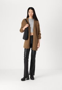 Cárdigan de punto marrón, top corto gris, pantalones de imitación cuero negros, botas de plataforma negras y un bolso de hombro negro. Fondo liso.