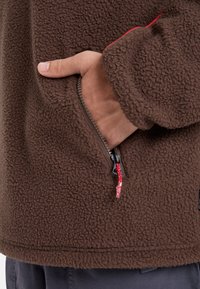 Chaqueta de forro polar marrón con tejido texturizado. Cuenta con un bolsillo con cremallera y tirador rojo, y un ajuste relajado. Primer plano de una mano en el bolsillo.