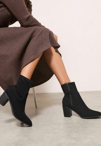 Černé semišové kotníkové boty s špičatou špičkou a blokovým podpatkem. Obsahují boční zip a texturovaný design látky.