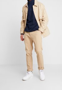 Giacca leggera beige sopra un pullover blu navale con mezza zip, abbinata a pantaloni beige chiaro e sneakers bianche. Linee pulite e trame semplici.