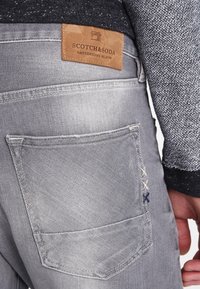 Harmaat farkut, joissa on nahkainen etiketti, jossa lukee "SCOTCH&SODA". Takataskussa on koristeompeleet, jotka lisäävät yksityiskohtia.