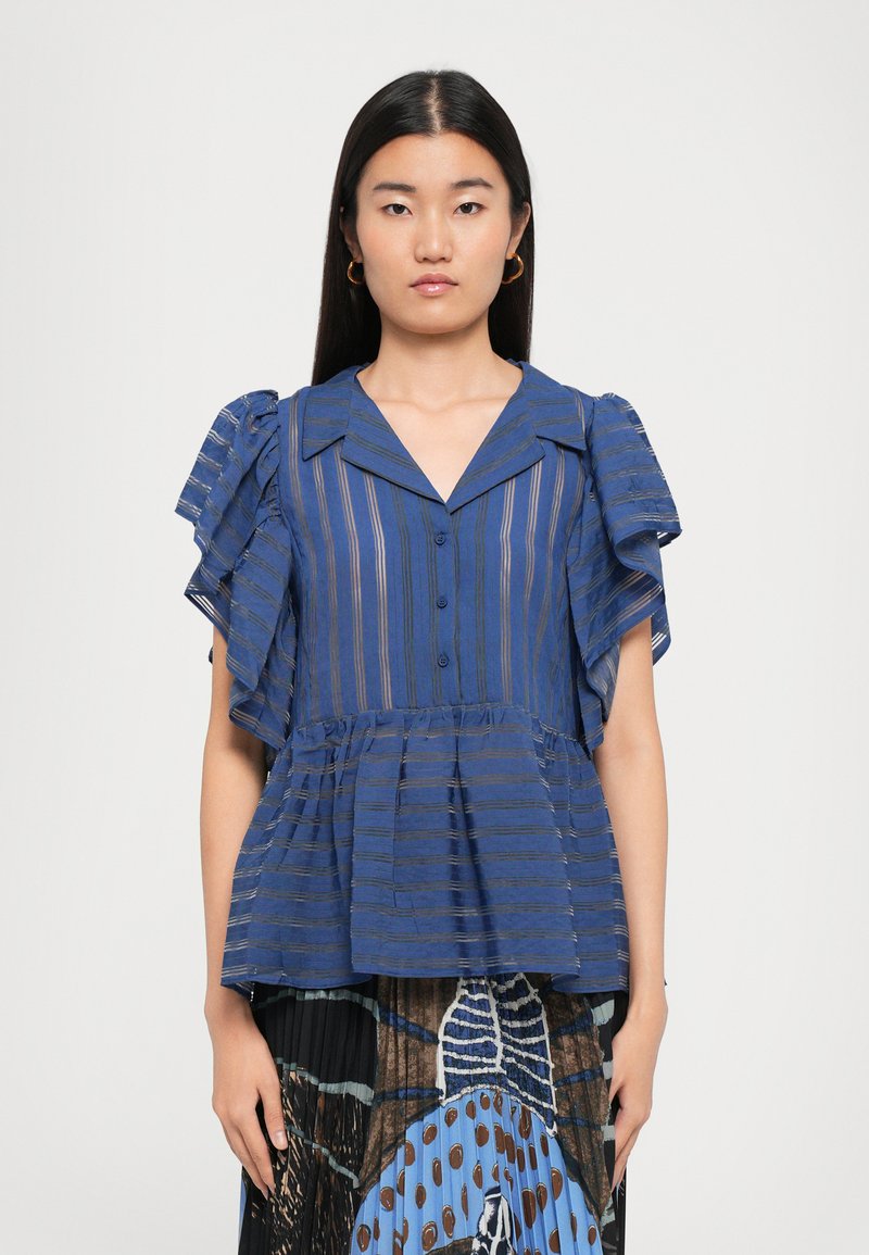 Woman with long dark hair wears blue striped blouse with ruffled sleeves and patterned pleated skirt, standing against plain background.