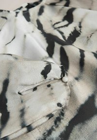 Hedvábná blůza s černo-krémovým abstraktním potiskem. Obsahuje malou kapsu s knoflíkovým uzávěrem a hladkou texturu.