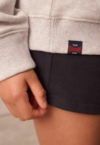 Šedá mikina s rebrovanými manžetami v kombinácii s čiernymi látkovými nohavicami. Na okraji je viditeľná červená a námornícka Superdry nálepka. Textúrované materiály.