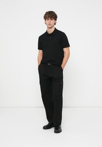 Svart poloshirt med smal passform og korte ermer, kombinert med svarte cargobukser med sidelommer. Modellen har på seg svarte sko.