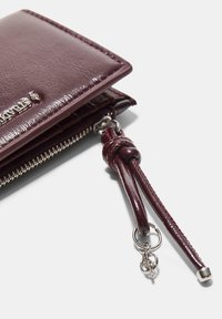 Portfel z bordowej skóry o fakturowanej powierzchni, wyposażony w zamek błyskawiczny, metalowe dodatki oraz dekoracyjny skórzany pasek z węzłem.
