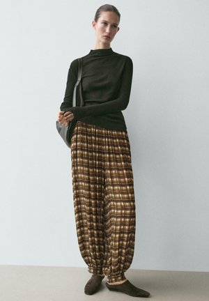 Donna in piedi contro un muro semplice, indossa una maglia nera a manica lunga, pantaloni ampi a quadri marroni, scarpe scure senza lacci e porta una borsa nera a tracolla.