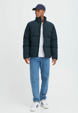 Schwarze Daunenjacke mit strukturiertem Schulterbereich, Frontreißverschluss und hohem Kragen. Kombiniert mit hellblauen Jeans und weißen Sneakers.