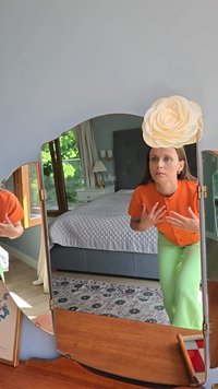 En tri-fold spegel med en rosaccent ovanpå reflekterar en person i en orange skjorta med knappar och gröna rutig byxor. En mönstrad matta är synlig.