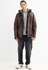 Chaqueta de cuero marrón con acentos acolchados en los hombros y capucha forrada de forro polar, usada sobre una camiseta gris y jeans negros, combinada con zapatillas blancas.