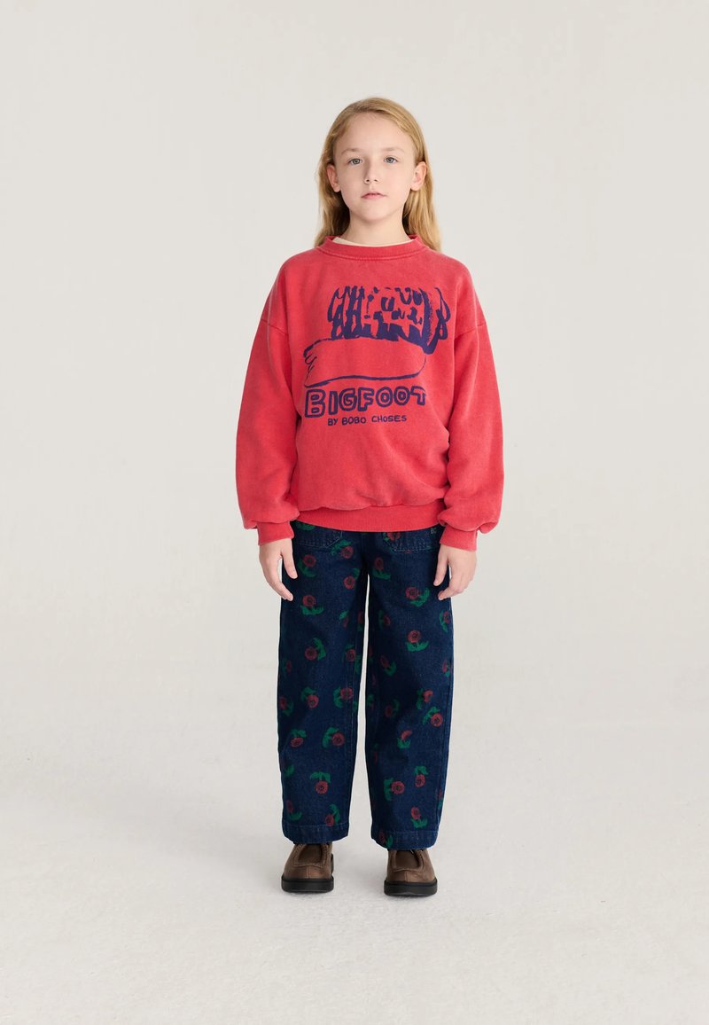 Enfant aux longs cheveux blonds se tenant face à nous, portant un sweat-shirt rouge avec le texte "Bigfoot" et un pantalon foncé avec des impressions florales rouges.