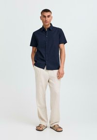 Camisa de botones de manga corta en color navy, fabricada en un material ligero, combinada con pantalones de lino beige claros y sandalias marrones, en una pose contra un fondo neutro.