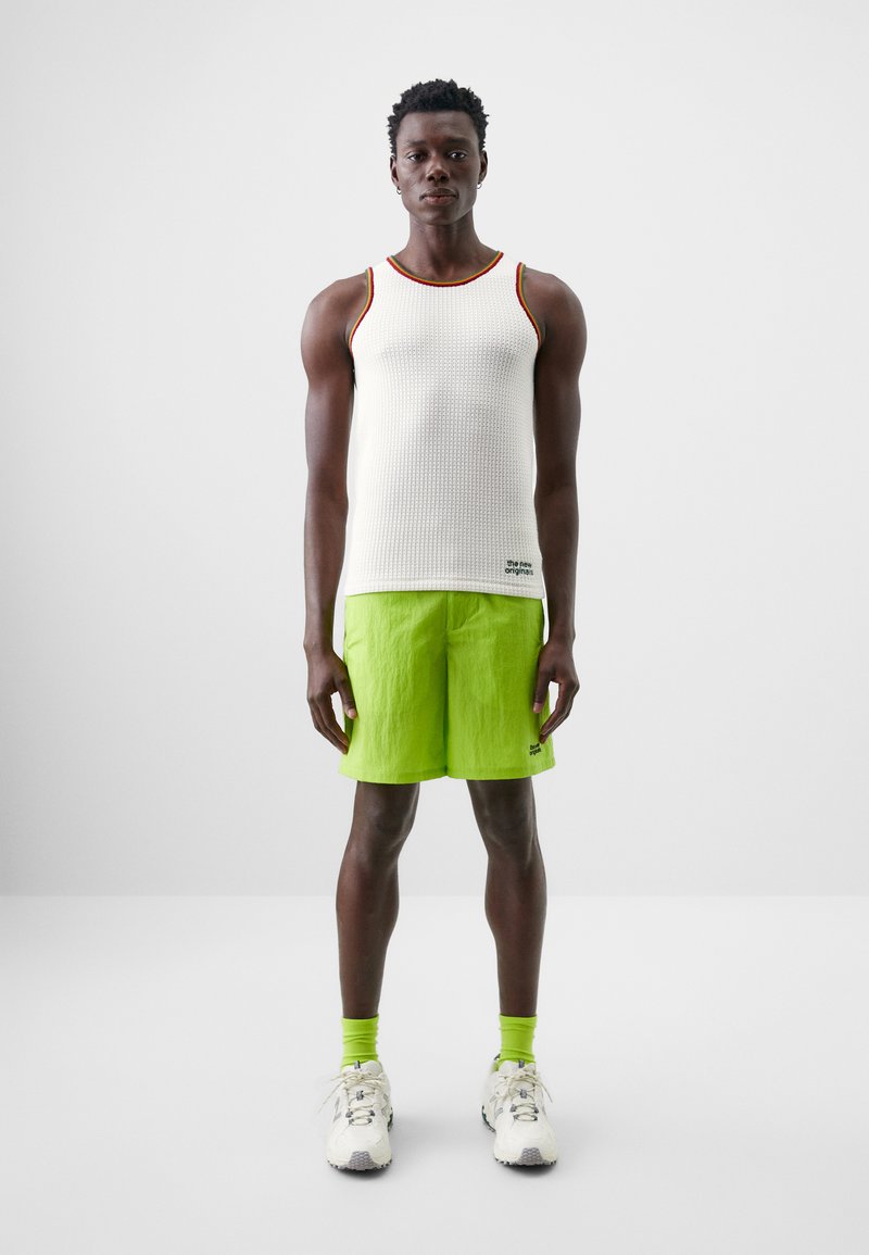 Hvit ribbet tanktop med rød og gul trim, kombinert med lysegrønne shorts. Merkbare detaljer inkluderer neongrønne sokker og hvite joggesko.