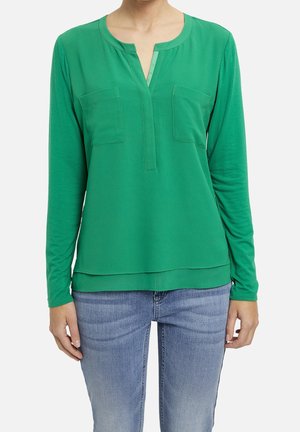 Kobieta ubrana w zieloną bluzkę z długim rękawem z dwoma przednimi kieszeniami na piersi oraz jasnoniebieskie dżinsy, stojąca na białym tle.