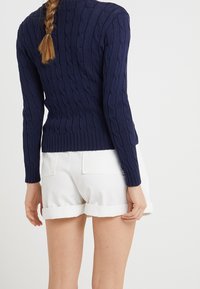 Vue arrière d'une personne portant un pull en maille bleu marine et un short blanc retroussé, avec des cheveux tressés pendants dans le dos.