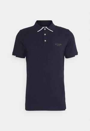 Granatowa koszulka polo z krótkim rękawem, białe wykończenia na kołnierzyku i rękawach, trzy guziki oraz logo "Guess EST.1981" na piersi.