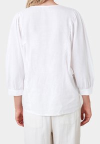 Blusa bianca a maniche lunghe con tessuto testurizzato, caratterizzata da un collo rotondo e maniche arricciate, indossata con pantaloni di colore chiaro abbinati.