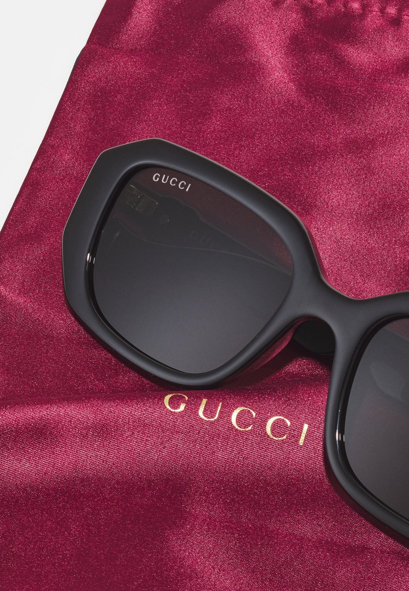 Schwarze Gucci-Sonnenbrille mit geometrischem Rahmen, präsentiert in einem bordeauxfarbenen Satinbeutel mit goldener Markenprägung. Glatte Textur und elegantes Design.