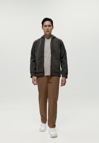 Tmavo šedá mikina na zips s dvoma prednými vreckami, v kombinácii so svetlým béžovým tričkom, hnedými nohavicami a bielymi teniskami. Hladká textúra, voľný strih.