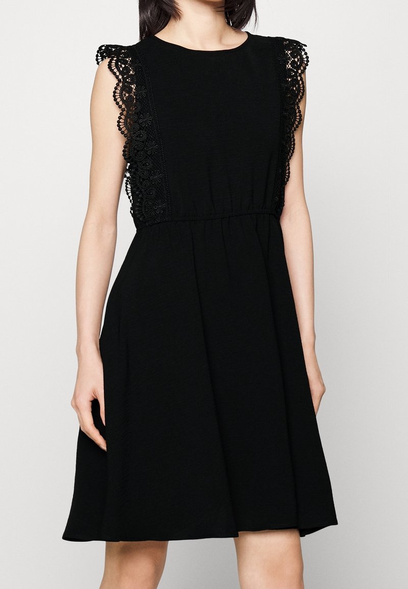 Femme portant une robe noire sans manches avec une bordure en dentelle florale sur les épaules et les côtés, taille cintrée et jupe longueur genou.
