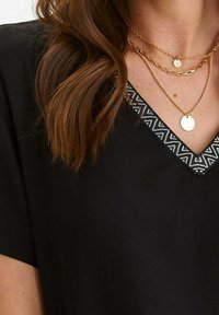 Černý top s výstřihem do V, který má lemování se zigzagovým vzorem. Zlaté vrstvené náhrdelníky s kruhovými přívěsky. Měkká textilie.