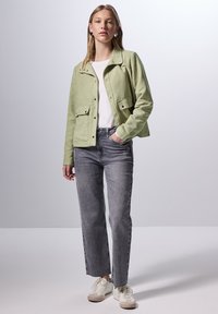 Chaqueta de ante verde claro con cierre de botones y dos bolsillos frontales, combinada con vaqueros rectos grises y zapatillas blancas. Diseño minimalista.