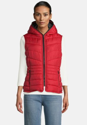 Vrouw in een rode gewatteerde mouwloze hoodie-gilet over een witte geribde lange mouw top en blauwe jeans, staand tegen een effen achtergrond.