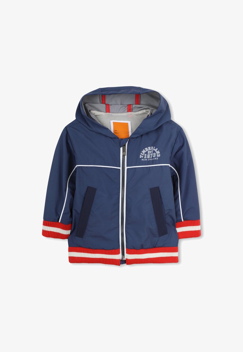 Veste coupe-vent bleu marine avec fermeture éclair avant, poignets rayés rouges et blancs, poches latérales, et graphisme "Timberland 1973" sur la poitrine.