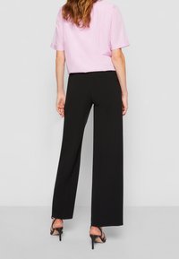 Femme avec de longs cheveux ondulés portant une chemise rose à manches courtes, un pantalon large noir et des sandales à talons hauts noires, de dos.