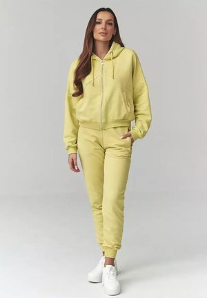 Kobieta stoi, ubrana w żółtą bluzę z zamkiem na suwak, dopasowane spodnie dresowe i białe sneakersy na platformie, na jednolitym szarym tle.