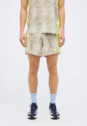 Άνδρας που φοράει μπεζ αθλητικά σορτς με σχέδια και ταιριαστό αμάνικο τοπ με νέον ρίγες, ανοιχτά μπλε κάλτσες και σκούρα μπλε αθλητικά παπούτσια.