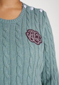 Ljusblått kabelstickat tröja med rund halsringning, som har tre dekorativa knappar och en vinröd broderad patch på bröstet.