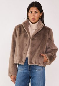 Veste courte marron en fausse fourrure avec col, fermeture par boutons et poches latérales. Portée sur un col roulé beige et un jean bleu.